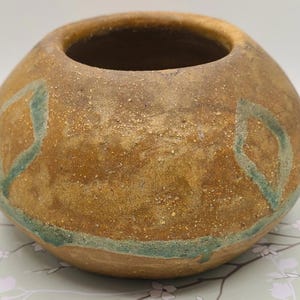 May include: A round, handcrafted ceramic vase with a wide opening. The vase has a textured, earthy brown exterior with accents of teal-colored diamond shapes. The interior is a darker brown.