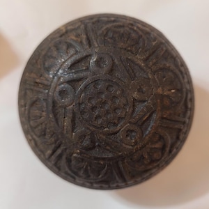 May include: A round, dark brown button with an intricate, embossed design. The button features a central circular pattern surrounded by stylized floral or geometric elements. The button's surface has a slightly aged or weathered appearance.