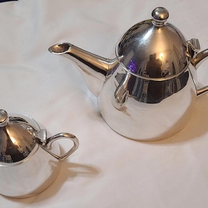 May include: A silver teapot and creamer set. The teapot has a rounded body and a lid with a knob. The creamer has a rounded body and a handle.