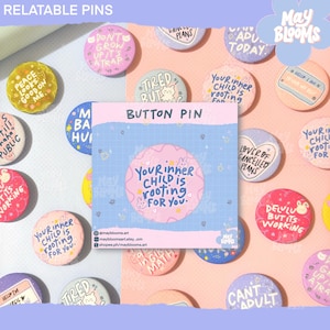 Pode incluir: Uma coleção de botões coloridos com vários designs e textos. Os botões apresentam frases como "Your inner child is rooting for you" e "Delulu but it's working." Os botões são redondos e têm um acabamento brilhante.