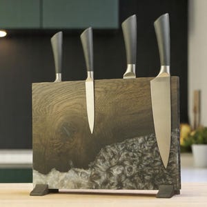Puede incluir: Un bloque de cuchillos de madera con cuatro cuchillos. El bloque está hecho de madera oscura y resina, con un patrón de vetas de madera natural. Los cuchillos tienen hojas plateadas y mangos negros. El bloque está sobre una superficie de madera.
