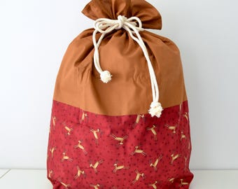 May include: A large, drawstring gift bag with a two-tone design. The top portion is a solid brown, while the bottom features a red fabric with a pattern of reindeer and stars. The bag is tied with a white rope.