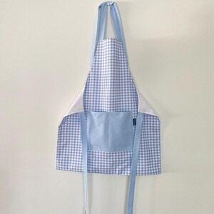 May include: A blue and white gingham apron with a light blue pocket and straps. The apron is hanging against a white wall. The apron is made of fabric.