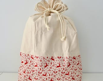 May include: A cream-colored drawstring gift bag with a red and white Christmas pattern at the bottom. The pattern features reindeer, snowflakes, and other festive elements. The bag is tied at the top with a cream-colored drawstring.