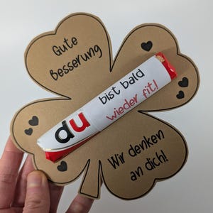 Peut inclure: Une carte de prompt rétablissement en forme de trèfle en papier brun, ornée de cœurs noirs et de texte allemand. Une barre chocolatée blanche et rouge est placée sur le trèfle, avec l'inscription "du bist bald wieder fit!"
