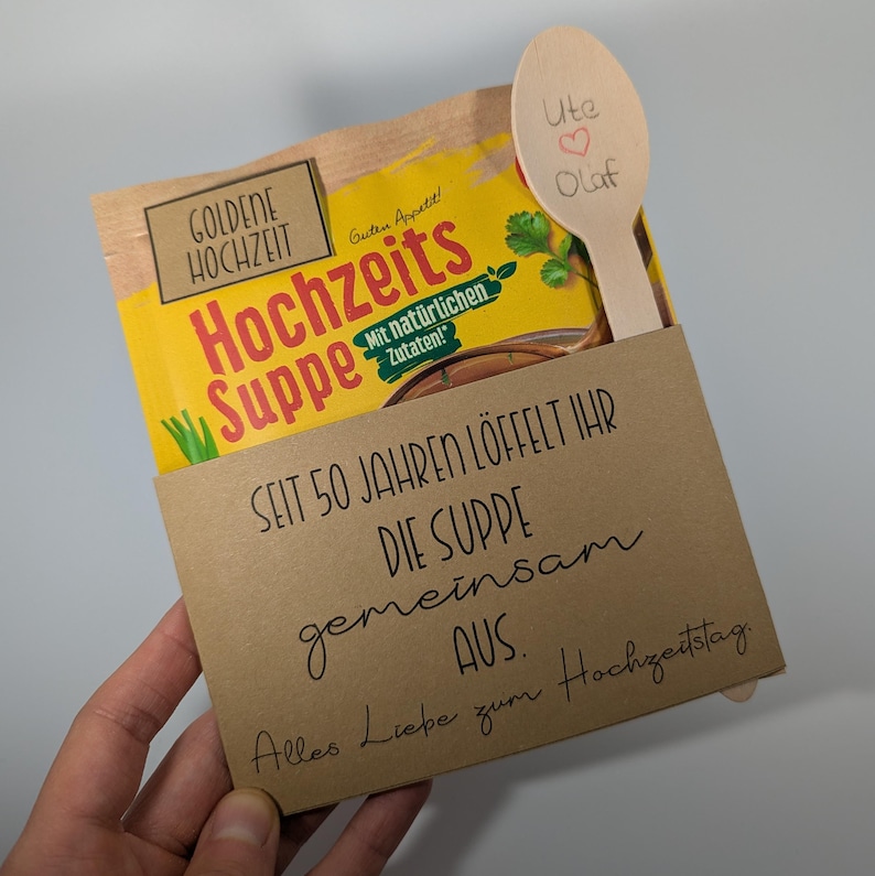 May include: A gift set featuring a packet of German soup, a wooden spoon, and a card. The soup packet is yellow with German text. The wooden spoon is engraved with names and a heart. The card has handwritten text.