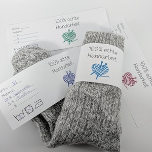 Puede incluir: Un par de calcetines grises de punto con una etiqueta blanca con el texto "100% echte Handarbeit" y un logotipo de ovillo de lana. Los calcetines son de talla 38, hechos de 95% algodón y 5% elastano. La etiqueta también incluye instrucciones de lavado.