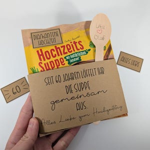 Op de afbeelding: Een cadeaupakket met een soepzakje, een houten lepel en een kaart. Het soepzakje is geel en bruin met Duitse tekst. De houten lepel heeft de namen "Ute" en "Olaf" met een hartje. De kaart luidt "Seit 60 Jahren löffelt ihr die Suppe gemeinsam aus."