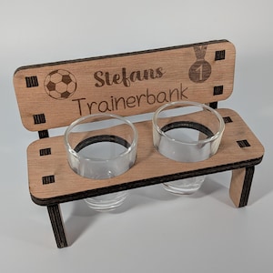 Peut inclure: Un objet en forme de banc en bois avec deux supports en verre transparent. Le banc porte l'inscription "Stefans Trainerbank" et un ballon de football. Un dessin de médaille avec le numéro "1" est également présent. L'objet est en bois brun clair.