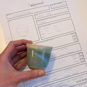Puede incluir: Una pequeña taza de espresso hecha a mano con un esmalte turquesa y crema. La taza se sostiene frente a una página de diario manuscrita con el título "Töpferjournal" y la palabra "Espressotasse". La altura de la taza es de 5,5 cm.