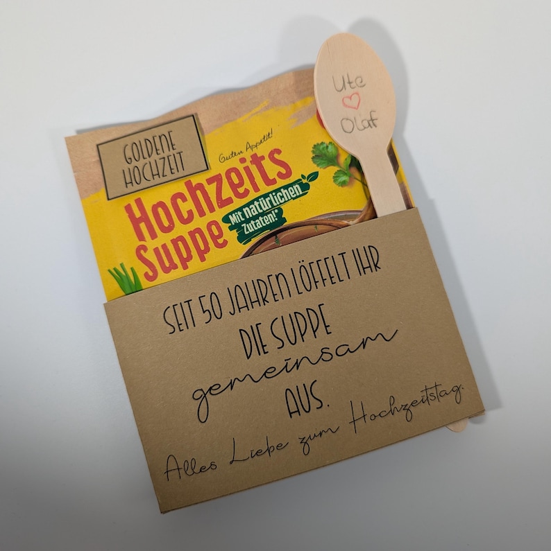 May include: A gift set featuring a package of "Hochzeits Suppe" soup, a wooden spoon with "Ute ♡ Olaf" written on it, and a brown card with German text. The soup package is yellow and brown.