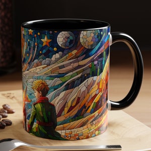 May include: A black ceramic mug with a colorful stained glass design featuring a character from The Little Prince. The design depicts a starry night sky with a crescent moon and a small planet.