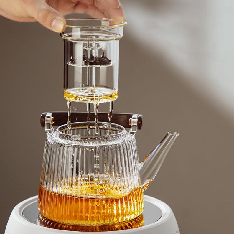 Pode incluir: Uma chaleira de vidro transparente com um design canelado e uma al&ccedil;a de madeira. A chaleira tem um infusor de vidro remov&iacute;vel com um filtro de malha met&aacute;lica. A chaleira est&aacute; cheia de um l&iacute;quido dourado.