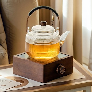 Może przedstawiać: Szklany czajnik z drewnianym uchwytem stoi na drewnianym elektrycznym podgrzewaczu do herbaty. Czajnik jest wypełniony jasnobrązową herbatą i ma żebrowany wzór. Podgrzewacz do herbaty ma okrągły pokrętło z przodu.