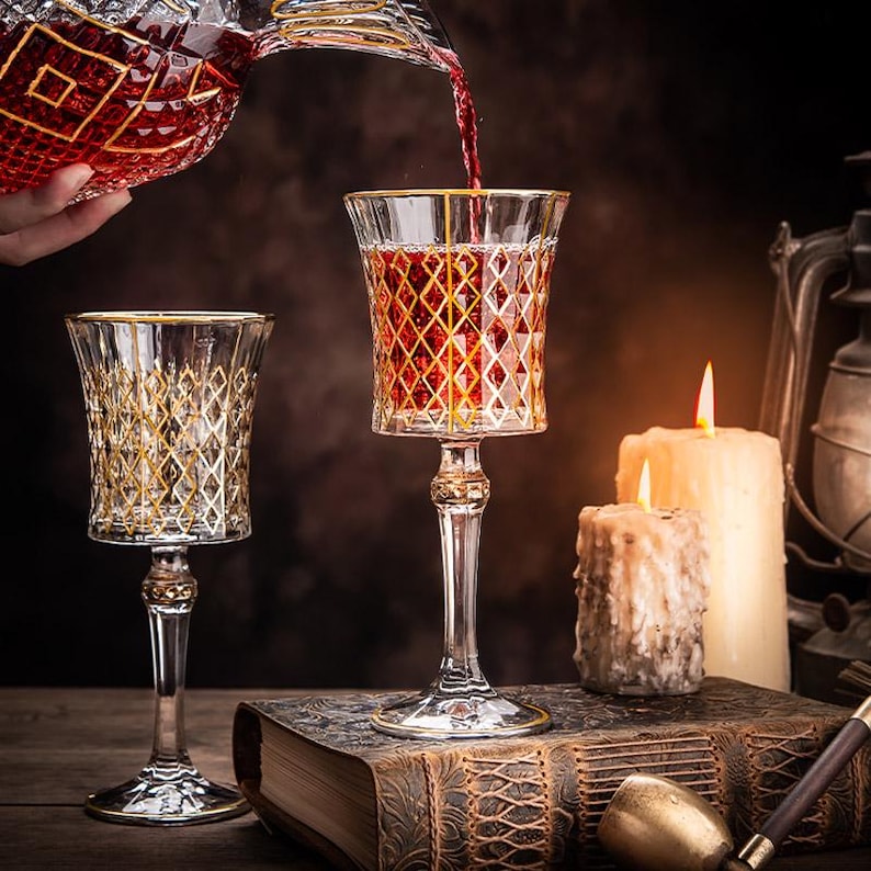 May include: Two wine glasses with gold rims and a geometric pattern, one filled with red wine, and a decanter pouring wine into the glass.