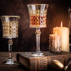 May include: Two stemmed glasses with gold rims and a diamond pattern. The glass on the right contains a red liquid. The glasses are on a book with a brown leather cover.