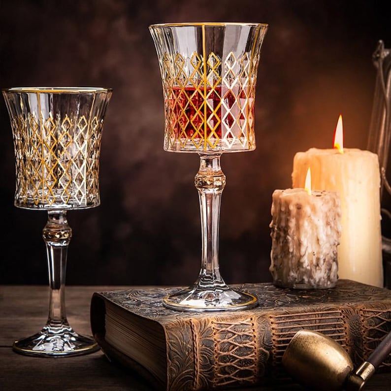 May include: Two stemmed glasses with gold rims and a diamond pattern. The glass on the right contains a red liquid. The glasses are on a book with a brown leather cover.