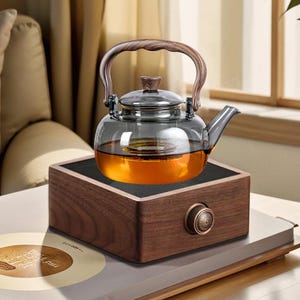 May include: A glass teapot with a wooden handle sits on a wooden electric tea warmer with a brass knob. The teapot is filled with amber-coloured tea.