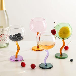 Puede incluir: Cuatro copas de vino coloridas con un diseño de tallo curvo único. Cada copa tiene una base y un tallo de diferente color, con un cuenco transparente. Una copa contiene arándanos, otra tiene un limón y una tercera tiene una cereza. Una botella de vino está en el fondo.