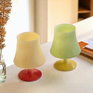 May include: Two glass goblets with a wide, flared bowl and a short stem. The goblet on the left has a cream-colored bowl and a red base. The goblet on the right has a light green bowl and a yellow base.