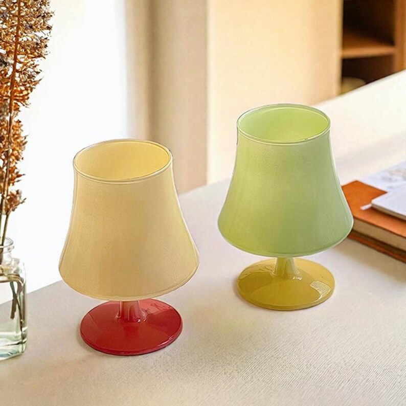 May include: Two glass goblets with a wide, flared bowl and a short stem. The goblet on the left has a cream-colored bowl and a red base. The goblet on the right has a light green bowl and a yellow base.