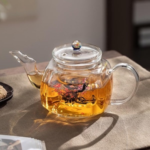 May include: A clear glass teapot with a floral design on the side. The teapot has a glass lid with a small knob on top. The teapot is filled with tea and is sitting on a brown surface.