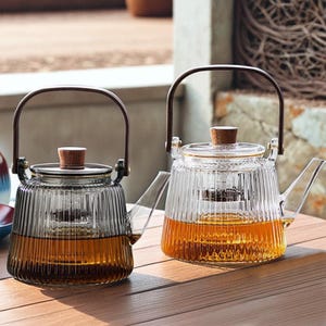 Puede incluir: Dos teteras de vidrio con asas y tapas de madera marrón. Las teteras tienen un cuerpo de vidrio transparente con nervaduras verticales y un filtro integrado. Las teteras están llenas de té marrón claro.