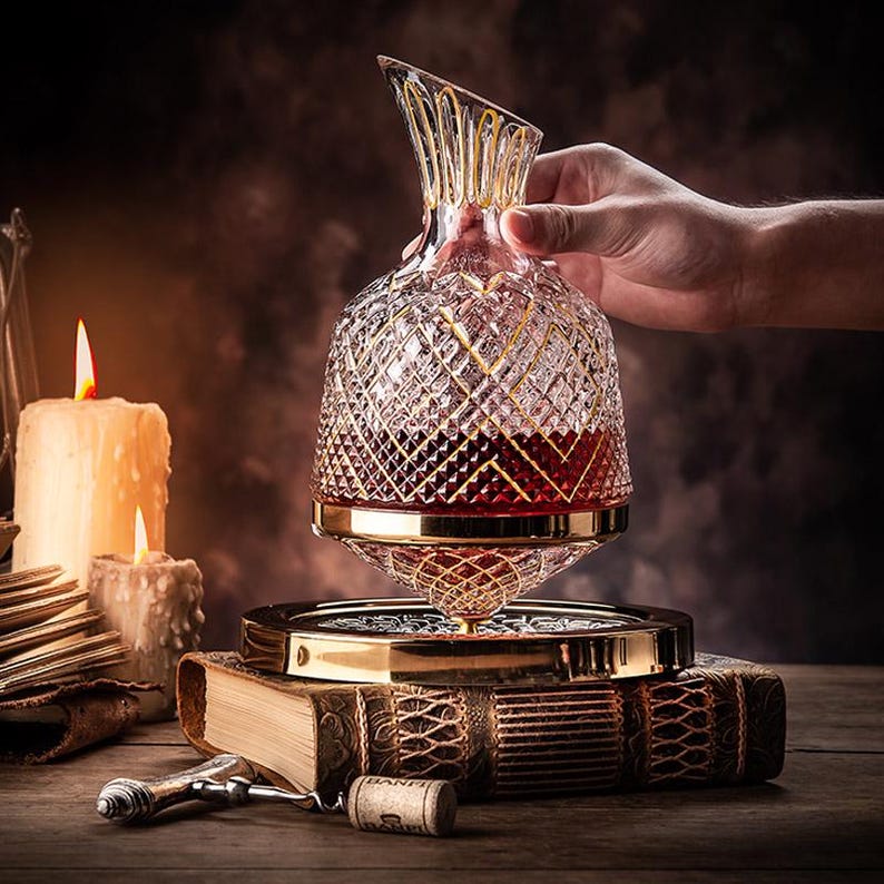 May include: A clear glass decanter with a gold-trimmed design, filled with red wine, sits on a gold-coloured stand. The decanter is being held by a hand, and the stand is resting on a book with a leather cover.