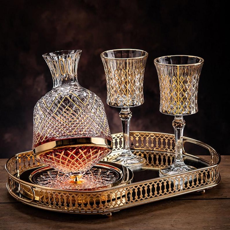 May include: A gold-trimmed glass decanter and two matching glasses sit on a gold-plated serving tray. The decanter and glasses have a diamond-shaped pattern.
