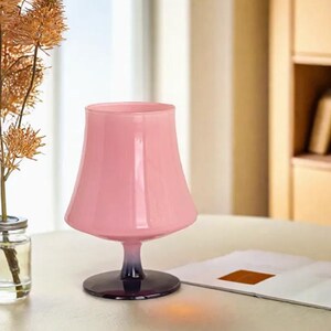 May include: A pink glass goblet with a wide bowl and a short stem. The goblet is sitting on a table with a book.