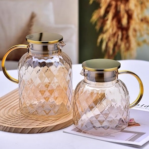 Puede incluir: Dos jarras de vidrio con tapas y asas doradas. Las jarras tienen un patrón de diamante y son transparentes con un ligero tono ámbar.