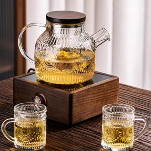 May include: A glass teapot with a wooden lid and handle, filled with a golden liquid, sits on a wooden base. Two matching glass mugs are in front. The teapot and mugs have a textured, patterned design, suggesting a tea set.