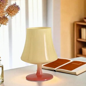 May include: A cream-colored glass table lamp with a wide, bell-shaped shade and a pink base. The lamp is sitting on a white table next to a book.