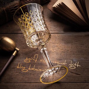 May include: A clear glass wine glass with a gold rim and intricate gold diamond pattern. The glass is sitting on a wooden surface.