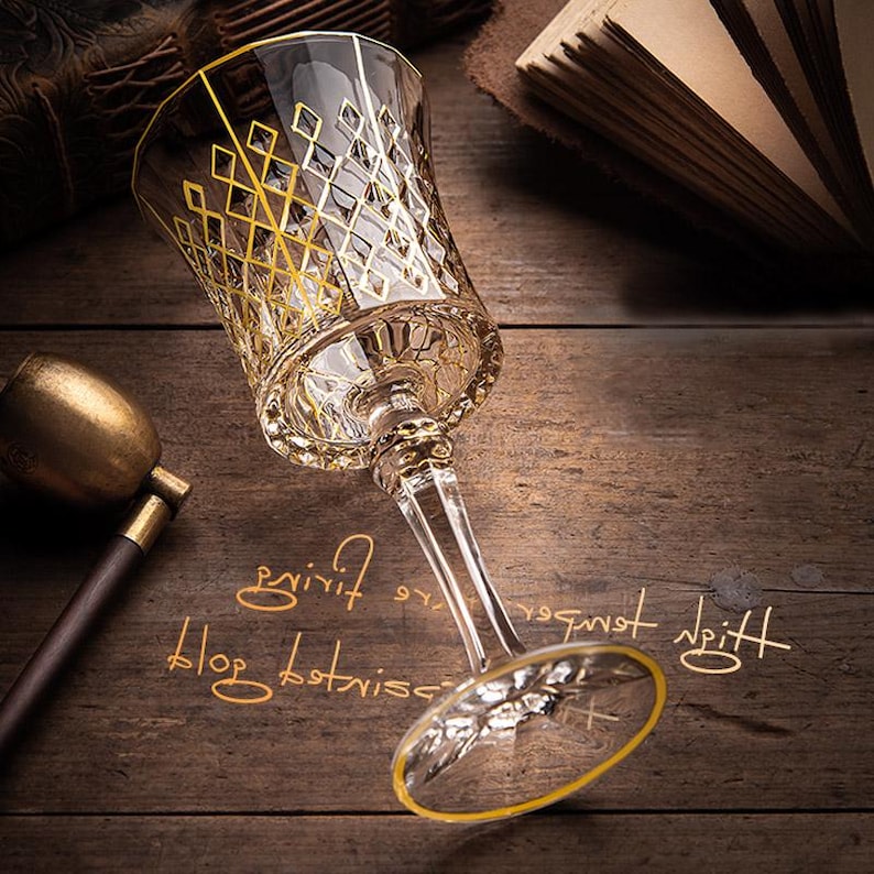 May include: A clear glass wine glass with a gold rim and intricate gold diamond pattern. The glass is sitting on a wooden surface.