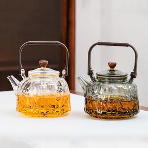 May include: Two glass teapots with brown handles and lids. One teapot contains a yellow liquid, the other a brown liquid. The teapots have a textured, ribbed design and are sitting on a white surface.