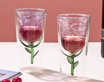 May include: Two clear glass cups with a rose design. The cups are filled with a red liquid, possibly wine. The roses are made of glass and have green stems and leaves.