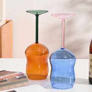以下が含まれることがあります： 鮮やかな色の装飾的なワイングラス2つ。1つは緑色のステムとトップが付いたオレンジ色、もう1つはピンク色のステムとトップが付いた青色です。グラスは丸みを帯びたベースと平らな円形のトップを備えています。