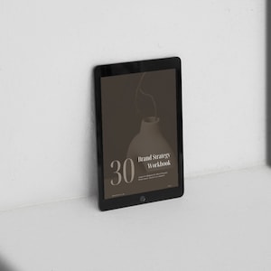 Może przedstawiać: Czarny tablet wyświetlający cyfrowy zeszyt ćwiczeń zatytułowany "Brand Strategy Workbook" z liczbą "30" w dużym białym foncie. Tło to brązowy wazon ze łodygą.