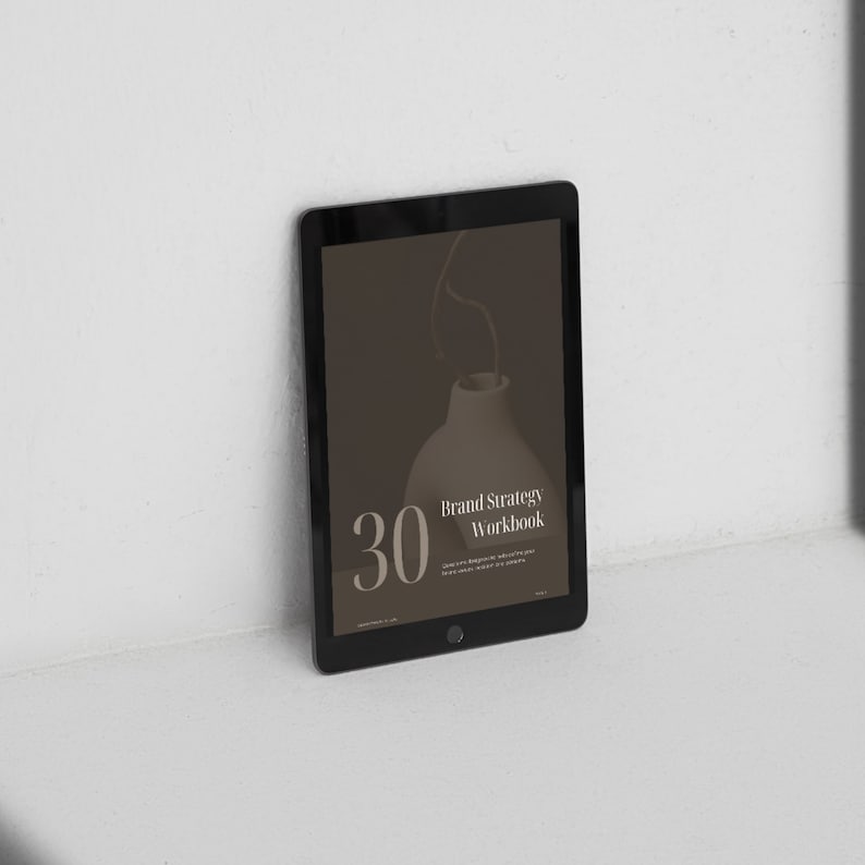 Może przedstawiać: Czarny tablet wyświetlający cyfrowy zeszyt ćwiczeń zatytułowany "Brand Strategy Workbook" z liczbą "30" w dużym białym foncie. Tło to brązowy wazon ze łodygą.