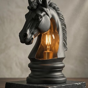 Puede incluir: Una lámpara de mesa negra mate con forma de pieza de ajedrez de cabeza de caballo. La sección hueca del cuello revela una bombilla estilo Edison de luz cálida sobre un interior dorado metálico. La escultura descansa sobre una base de piedra negra.