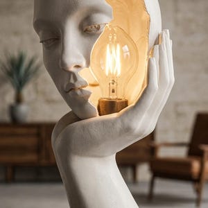 Puede incluir: Una lámpara de mesa escultural blanca que presenta un rostro humano con un lado agrietado que revela una bombilla tipo Edison brillante. La escultura muestra una mano acunando el rostro, con un acabado mate y luz cálida desde la cavidad.