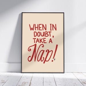 May include: A framed art print with the text "WHEN IN DOUBT, TAKE A Nap!" in red script on a cream background. The black frame is rectangular and the print is displayed against a white wall and a wooden floor.