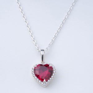 May include: A silver necklace with a heart-shaped pendant. The pendant has a large, faceted, ruby-red stone surrounded by a halo of small, clear gemstones. The chain is delicate and silver, set against a white background.