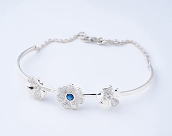 Pode incluir: Pulseira de prata com três pingentes em forma de flor. A flor central tem uma pedra azul, as outras são adornadas com pequenas pedras transparentes. Fecho de corrente delicado. Fundo branco.