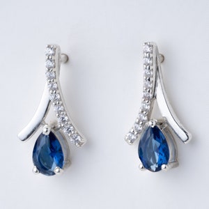 Op de afbeelding: Een paar zilveren oorbellen met een druppelvormige, diepblauwe edelsteen. De oorbellen hebben een gebogen ontwerp met een rij kleine, heldere edelstenen. De oorbellen staan tegen een witte achtergrond.