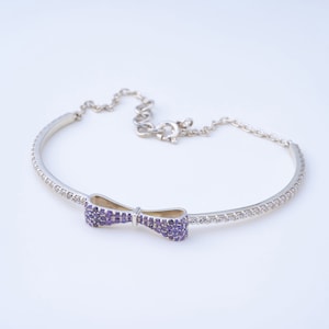 Puede incluir: Una pulsera de plata con un centro en forma de lazo. El lazo está adornado con pequeñas piedras preciosas moradas. La banda de la pulsera está adornada con piedras claras y brillantes. El cierre es una cadena con un cierre de mosquetón.