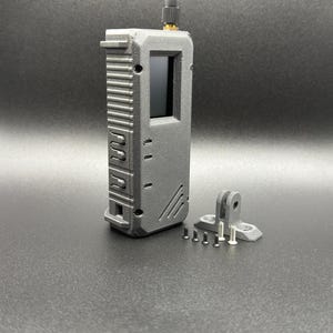 Könnte beinhalten: Ein schwarzes, rechteckiges Gerät mit einem kleinen Bildschirm und einer Antenne. Es hat eine strukturierte Oberfläche und eine kleine, schwarze Plastikhalterung mit Schrauben.