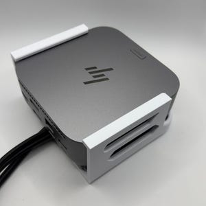 Könnte beinhalten: Ein dunkelgrauer Computer mit dem HP-Logo, montiert in einer weißen Halterung. Schwarze Kabel gehen von der Seite ab. Der Computer ist rechteckig mit abgerundeten Ecken. Die Halterung hat einen Schlitz, in den der Computer geschoben werden kann.