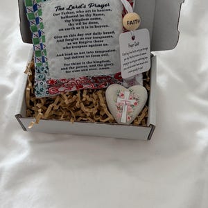 May include: A white cardboard gift box containing a small fabric square with a green and red patterned border, printed with The Lord's Prayer. A wooden bead labelled FAITH, a white tag labelled Prayer Quilt, and a heart-shaped stone with a floral cross.